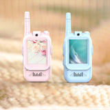 Two toy walkie-talkies, one pink and one blue, on a blurred natural background.