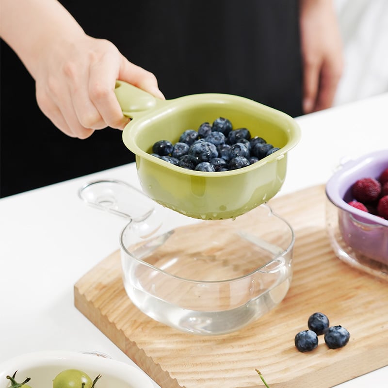 Person using a green colander to drain blueberries over a glass container on a wooden cutting board.